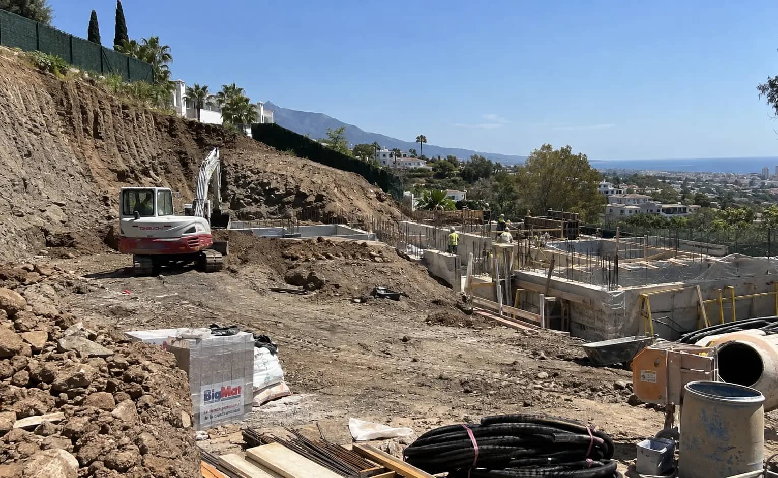 Hillside villa plot during groundworks, with foundation walls formed and a digger on site